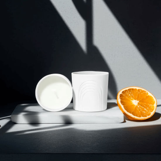 Komposition auf weißem Marmor: zwei weiße Kerzenglaser (eines stehend Bogenmuster, eines liegend weißes Wachs). Rechts eine saftige halbe Orange. Dramatische geometrische Schatten durchziehen die Szene, erzeugen starken Kontrast.
